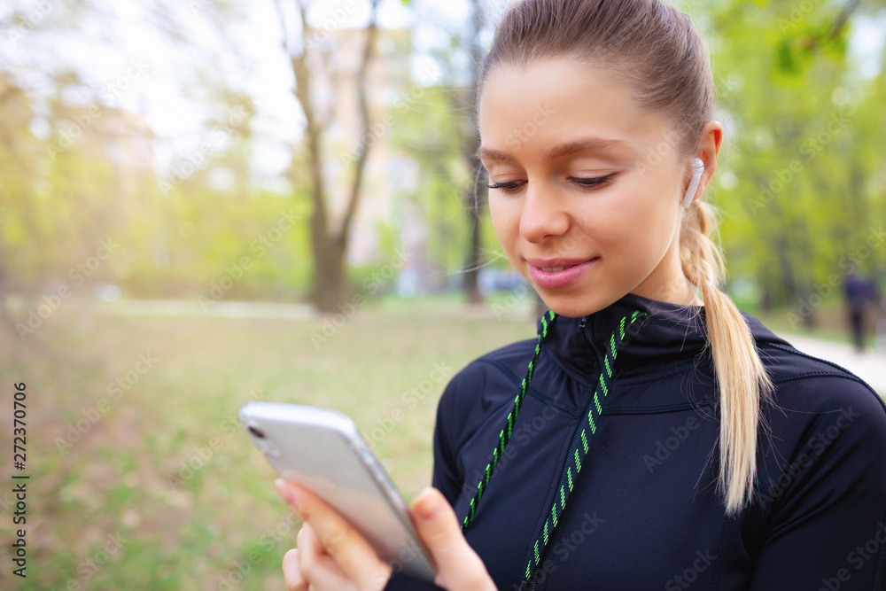 Young smiling girl making sport and running in the park using her phone to listen the music with wireless headphones on sunset in the city watching the screen and having communication talking chat Young smiling girl making sport and running in the park using her phone to listen the music with wireless headphones on sunset in the city watching the screen and having communication talking chat