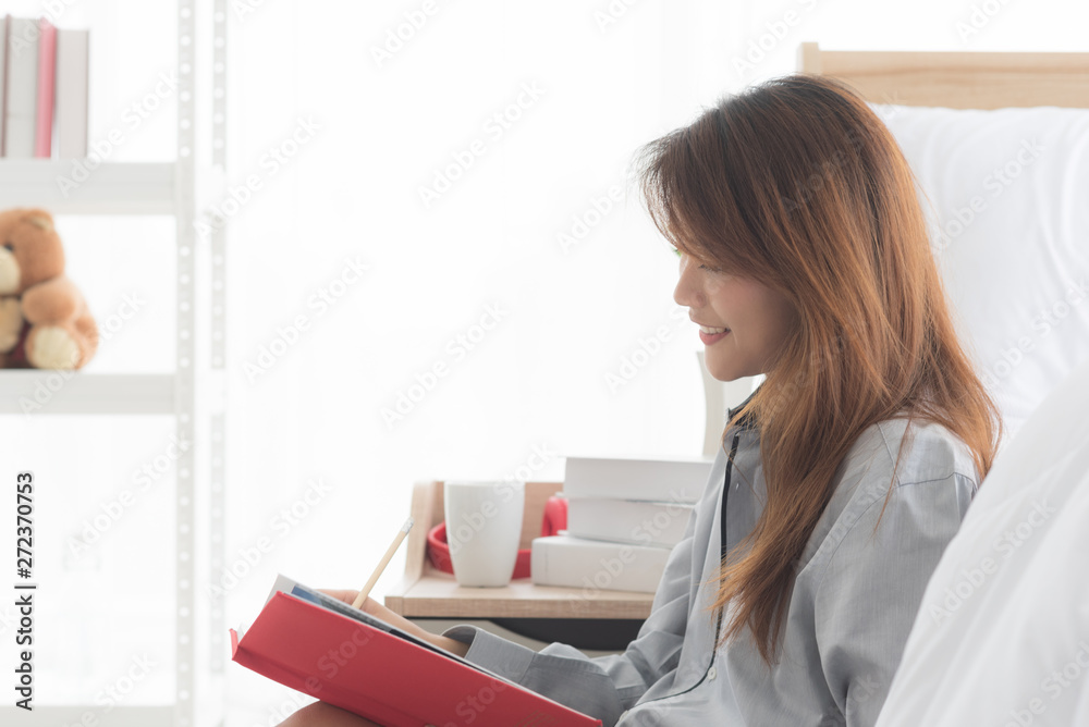 Young Asian smiling woman sitting beside bed and writing diary in modern bedroom Young Asian smiling woman sitting beside bed and writing diary in modern bedroom