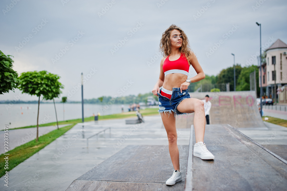 Sexy curly model girl in red top, jeans denim shorts, eyeglasses and sneakers posed at skatepark.