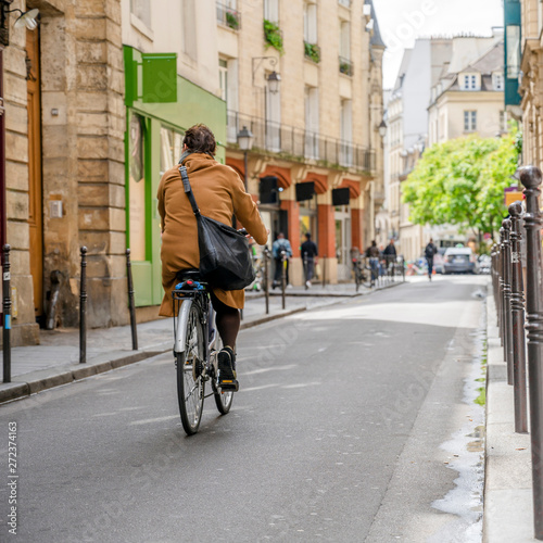 Woman with bag over her shoulder is riding bicycle along the street of old Paris with residential buildings and small shops
