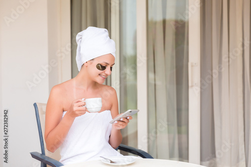 Happy smiling woman in under-eye patches readind sms message and drinking coffee at hotel terrace resort. morning holiday concept. Good news photo