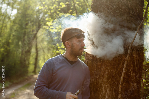 Vape man. An adult white bearded man in sunglasses smokes an electronic cigarette outside in the forest in sunny day. Bad habit that is harmful to health.