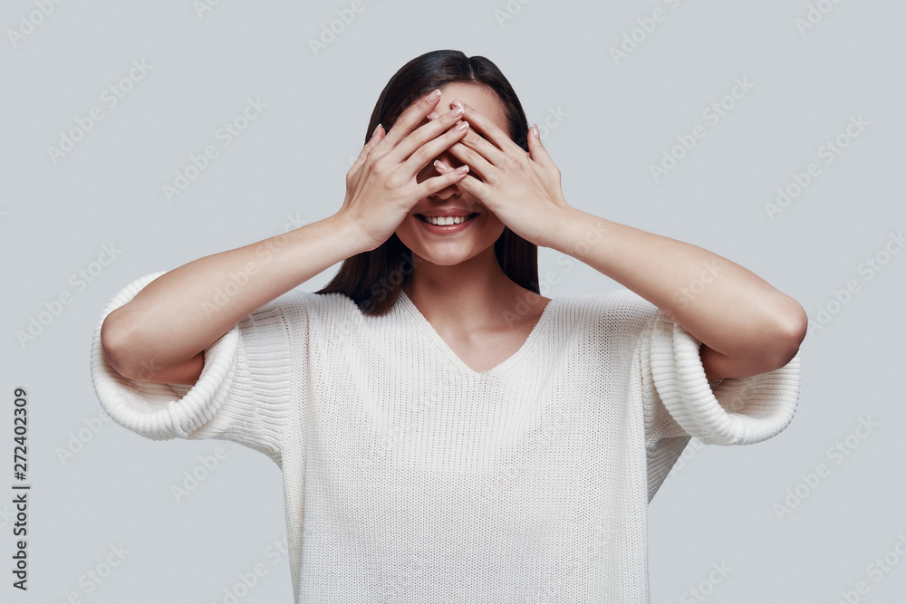 See nothing. Attractive young woman covering eyes with hands and smiling while standing against grey background See nothing. Attractive young woman covering eyes with hands and smiling while standing against grey background