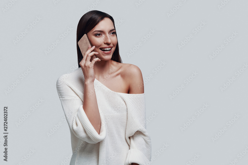 Pleasant talk. Attractive young woman talking on the smart phone and smiling while standing against grey background Pleasant talk. Attractive young woman talking on the smart phone and smiling while standing against grey background