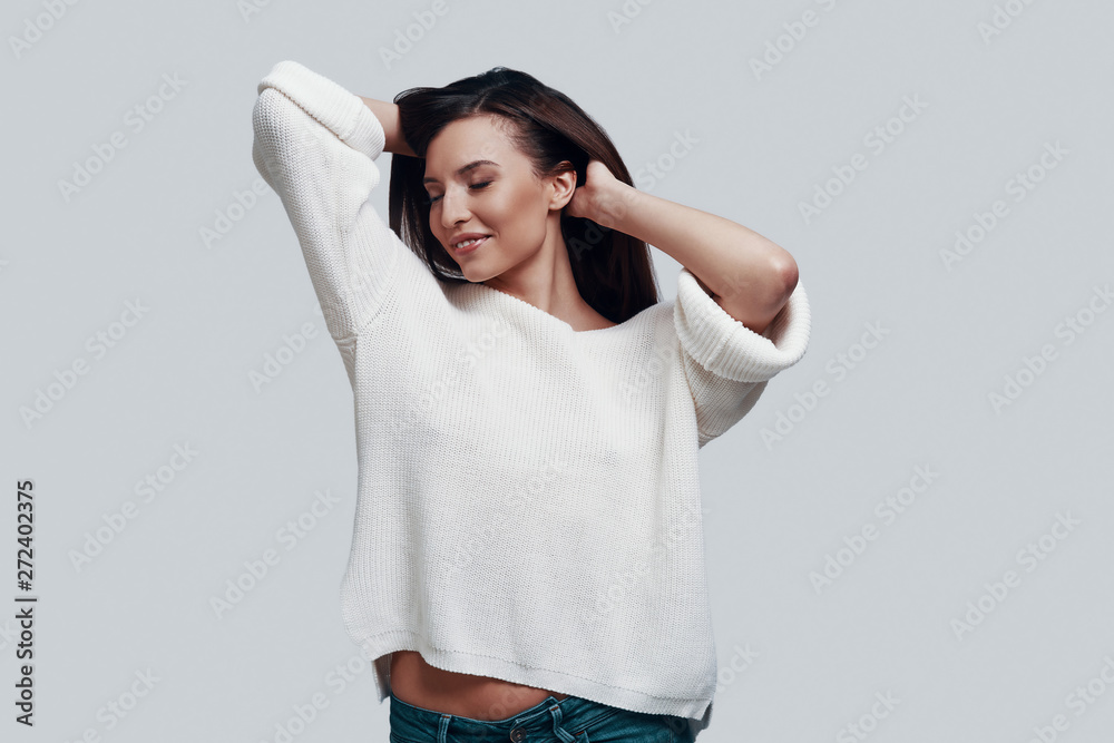 Pure feminine beauty. Attractive young woman keeping hands in hair and smiling while standing against grey background Pure feminine beauty. Attractive young woman keeping hands in hair and smiling while standing against grey background