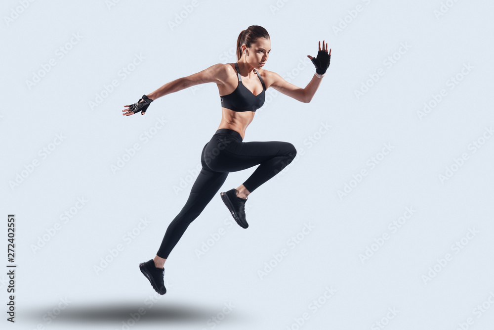 Challenging herself. Full length of attractive young woman in sports clothing jumping while hovering against grey background Challenging herself. Full length of attractive young woman in sports clothing jumping while hovering against grey background