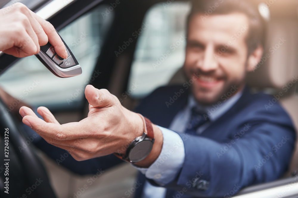 Thankful businessman. Blurred image of successful and happy young man in full suit is taking a key from his car and smiling while sitting on the front seat.