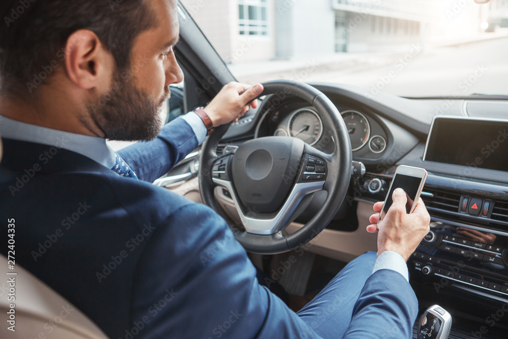 Confident businessman. Back view of confident and young bearded man in a stylish suit touching his smart phone while sitting in the car Confident businessman. Back view of confident and young bearded man in a stylish suit touching his smart phone while sitting in the car