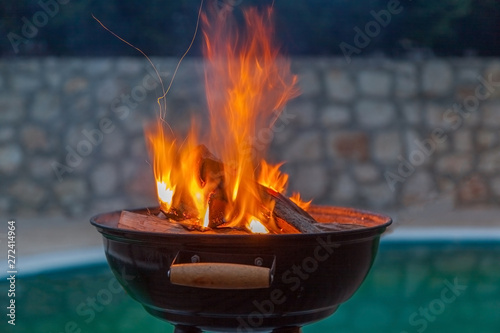 Round black grill on backyard is full of coal and wood in orange fire flames ready to cook with pool on background on summer sunset time, almost dark outside