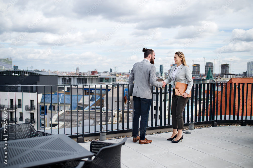 Two young business people standing on a terrace outside office, shaking hands.
