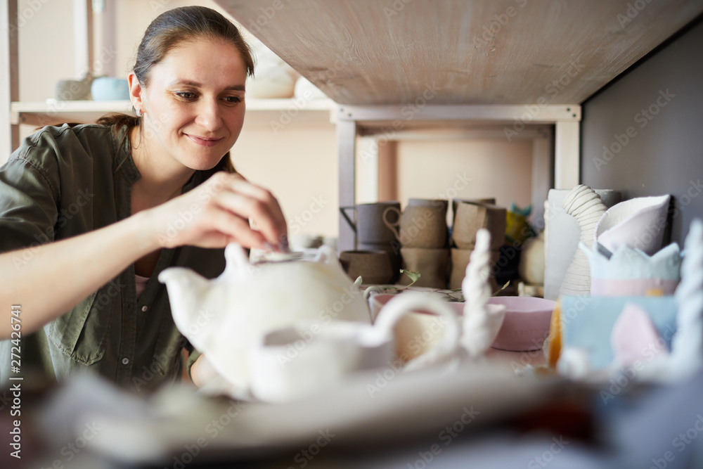 Portrait of contemporary female potter putting handmade kettle on shelf in studio, copy space Portrait of contemporary female potter putting handmade kettle on shelf in studio, copy space
