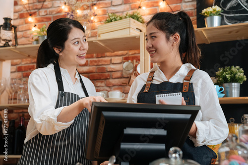 Team work and coworkers concept. two smiling female barista working together behind bar counter in coffeehouse. young girls coffee store staffs wear aprons using digital tablet talking and laughing.