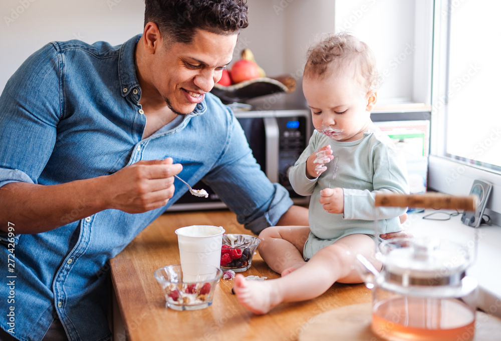 A father and a small toddler son eating fruit and yoghurt indoors at home.