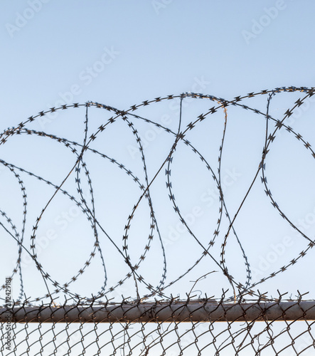 Tangled razor wire on top of a wire mesh perimeter fence, against a blue sky