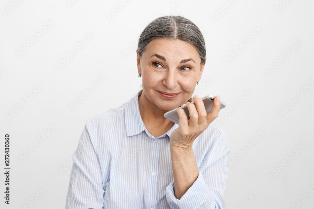 Beautiful elegant female manager in her sixties having playful look while using mobile at office, recording voice message. Attractive cheerful middle aged woman chatting online via smart phone Beautiful elegant female manager in her sixties having playful look while using mobile at office, recording voice message. Attractive cheerful middle aged woman chatting online via smart phone