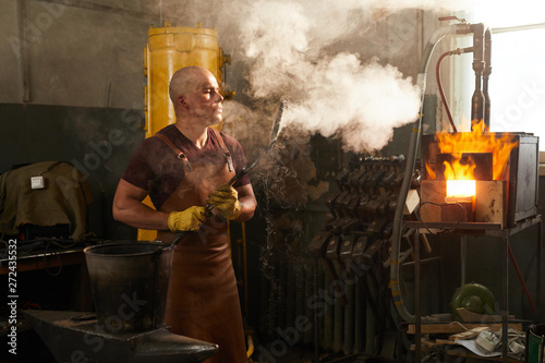 Foto Serious bald young male metalworker in leather apron blowing on forged knife in