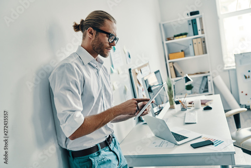 Cutting edge technologies. Good looking young man in shirt using digital tablet while standing in the office