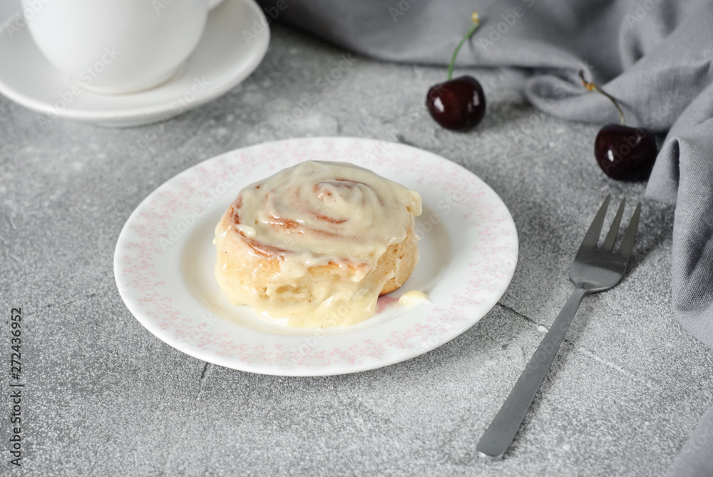 Cinnamon bun on a round plate on a gray background, decorated with a linen towel and cherry. Horizontal image.