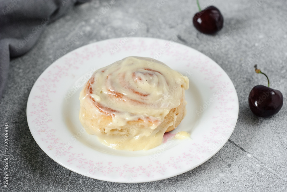 Cinnamon bun on a round plate on a gray background, decorated with a linen towel and cherry. Horizontal image.