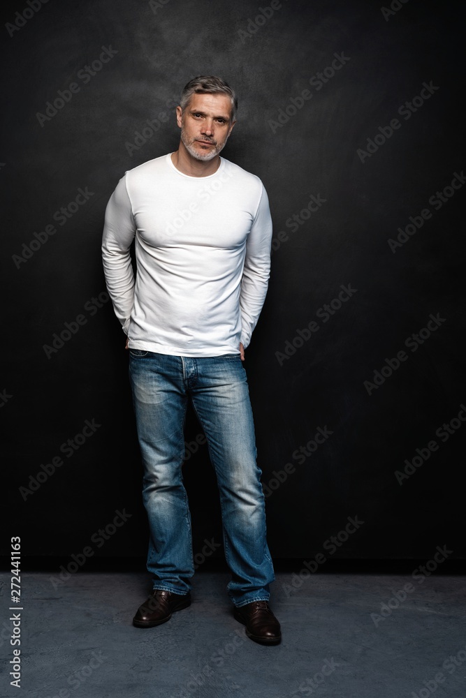 Full body portrait of middle-aged good looking man in white t-shirt posing in front of a black background with copy space.