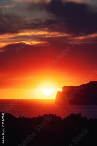 Dramatic ocean sunset scene with giant big sun setting behind a rock coastline silhouette an colorful sky glow. Port Andratx  Mallorca  Spain