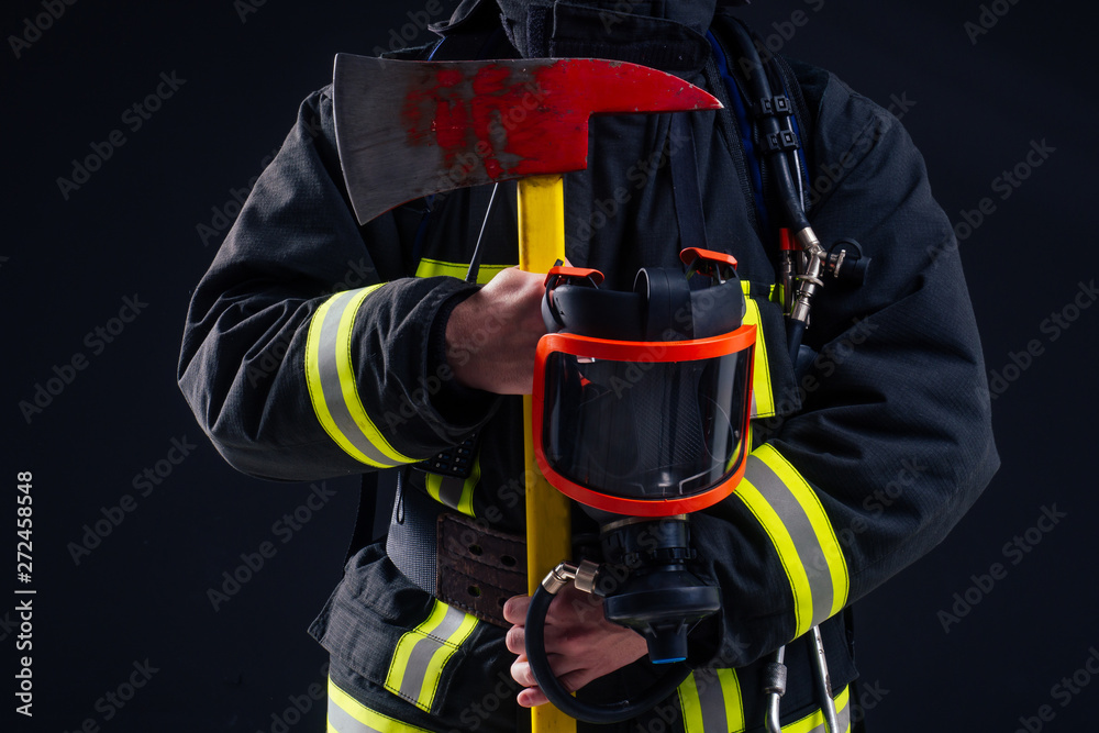 Fototapeta premium portrait strong fireman in fireproof uniform holding an ax chainsaw in his hands black background studio