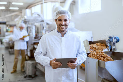 Young smiling manager in sterile uniform holding tablet and looking at camera while standing in food factory.