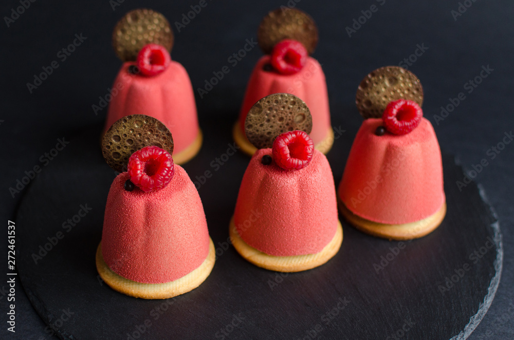 Set of chocolate mini mousse cakes covered with pink velvet spray, decorated with chocolate elements and fresh raspberry on black background