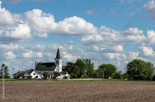 Beautiful, white church in a country, rural setting surrounded by farm fields and trees. Bright blue sky with puffy clouds create a beautiful landscape