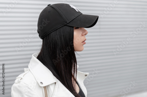 Canvas Print Portrait of pretty young beautiful woman in a stylish white leather jacket in a fashionable black baseball cap near a metal wall in the city