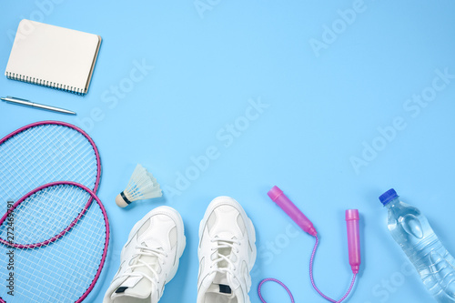 Sports flat lay with shuttlecock and badminton racket, skipping rope, sneakers, bottle water, pen and notepad on blue background. Fitness, sport and healthy lifestyle concept.