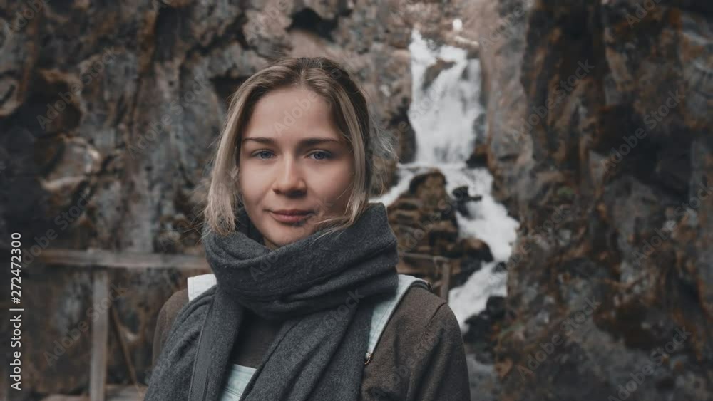 Close-up portrait: a beautiful young blonde girl in a scarf stands on the background of a waterfall, looking at the camera and smiling. Girl in a hike to a wild waterfall in the rocks. Slow motion, 4K Close-up portrait: a beautiful young blonde girl in a scarf stands on the background of a waterfall, looking at the camera and smiling. Girl in a hike to a wild waterfall in the rocks. Slow motion, 4K
