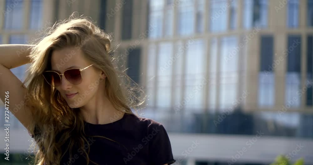 pretty young woman with trendy sunglasses on face is walking in city in summer, posing for camera pretty young woman with trendy sunglasses on face is walking in city in summer, posing for camera