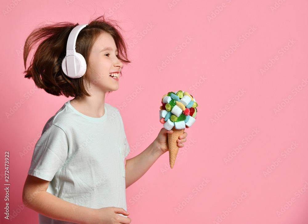 Happy teen girl kid walks in headphones sings with a big ice cream in waffles cone with tasty toppings give it on pink Happy teen girl kid walks in headphones sings with a big ice cream in waffles cone with tasty toppings give it on pink