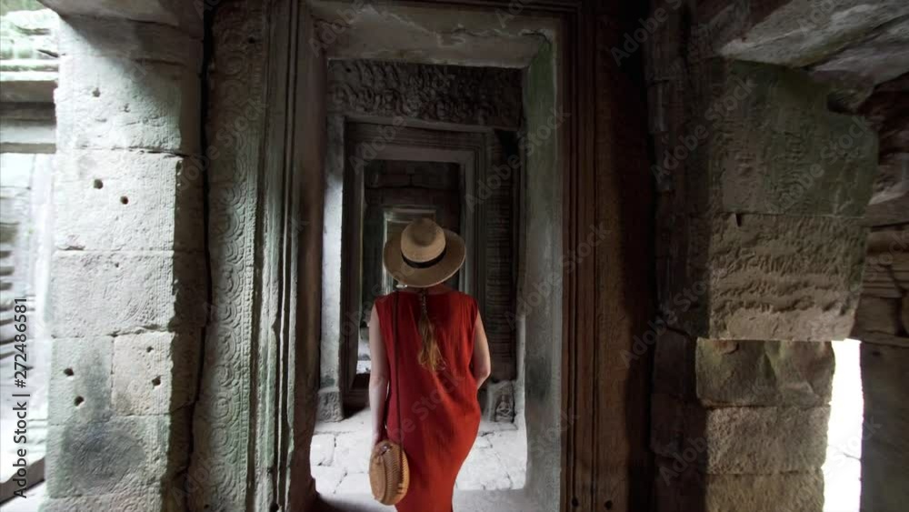 Caucasian woman in red dress is walking along narrow corridor of Bayon temple exploring it with interest. Angkor Wat complex, Cambodia Caucasian woman in red dress is walking along narrow corridor of Bayon temple exploring it with interest. Angkor Wat complex, Cambodia