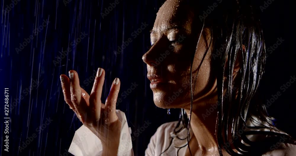 young brunette is getting wet under rain in night outdoors, holding palm up and looking to heaven