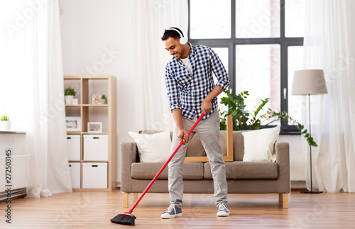 cleaning, housework and housekeeping concept - indian man in headphones with broom sweeping floor at home