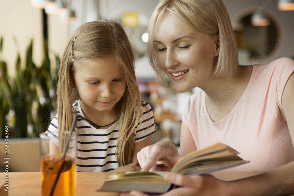 Front view of cheerful woman reading book to little daughter in cafe. Beautiful girl drinking juice and listening to mother. Cute family laughing and relaxing together. Concept of family time. Front view of cheerful woman reading book to little daughter in cafe. Beautiful girl drinking juice and listening to mother. Cute family laughing and relaxing together. Concept of family time.