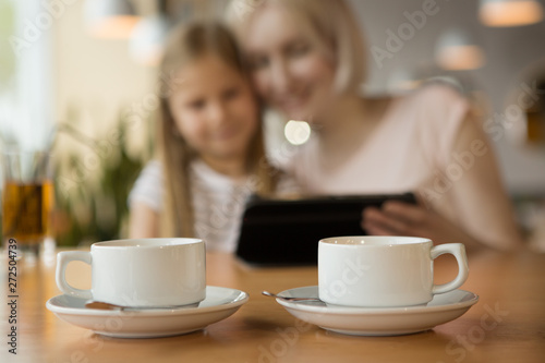 Two white cups of warm tea standing on table in cafe while mother and daughter sitting and watching video at background. Smiling woman and girl ordering cappuccino and coffee. Concept of lunch time.
