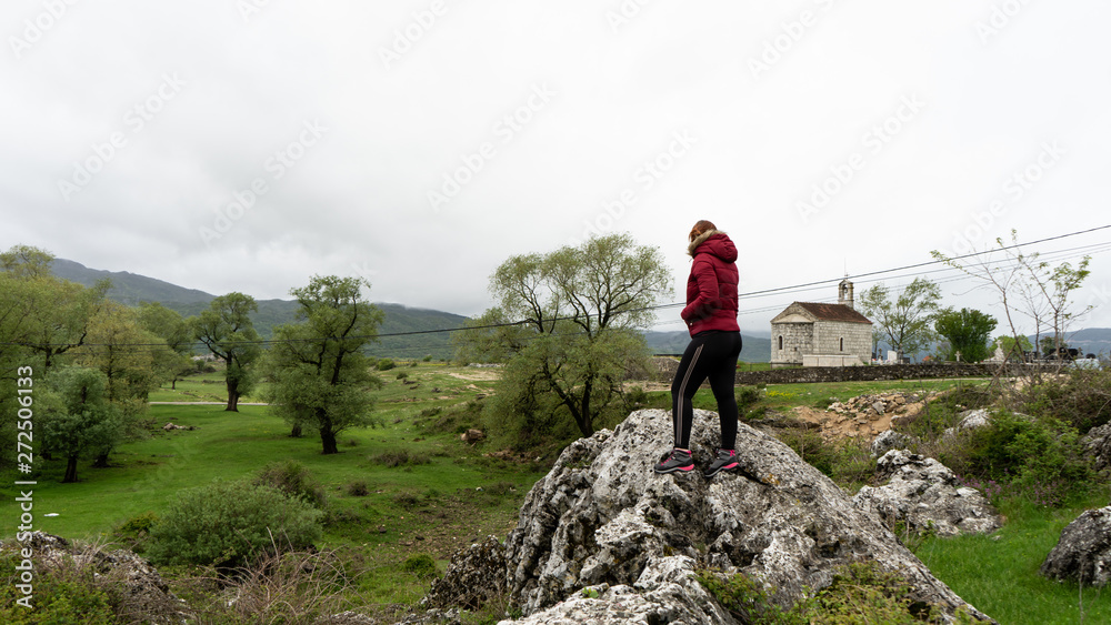 Girl in sport clothes and red winter jacket in a small village of Montenegro with small orthodox chapel