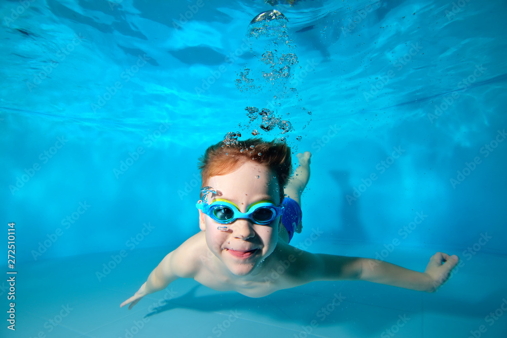 Happy smiling baby, boy, swimming underwater in swimming pool in swimming glasses and posing for camera on blue background. Portrait. Underwater photography. Vertical orientation