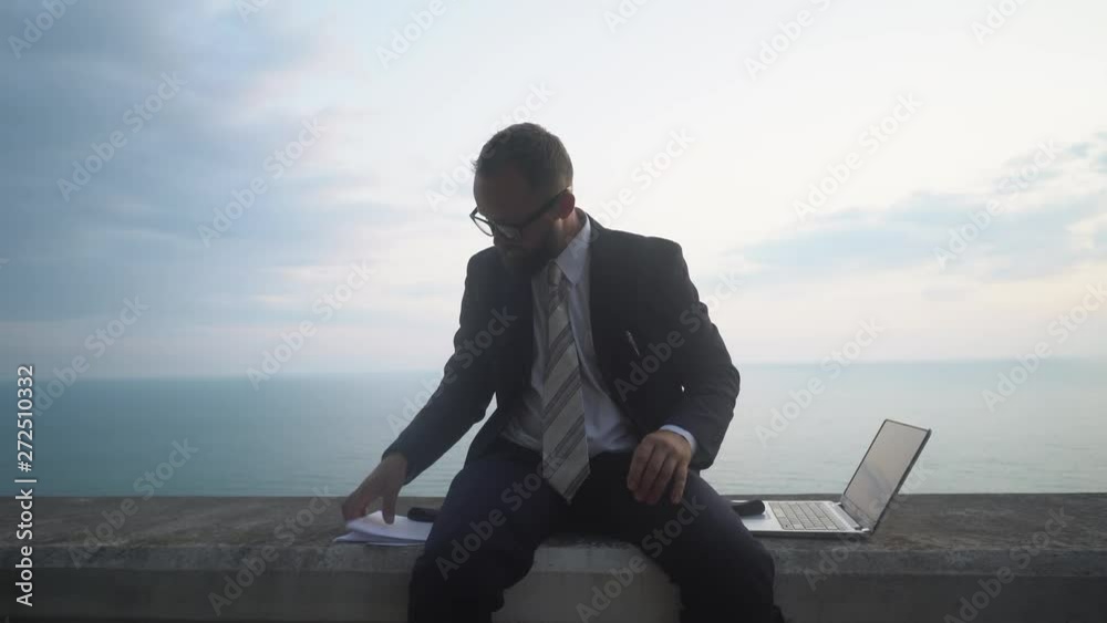 young businessman in a suit without shoes is sitting and working in a laptop on a concrete parapet on the background of a sea sunset young businessman in a suit without shoes is sitting and working in a laptop on a concrete parapet on the background of a sea sunset