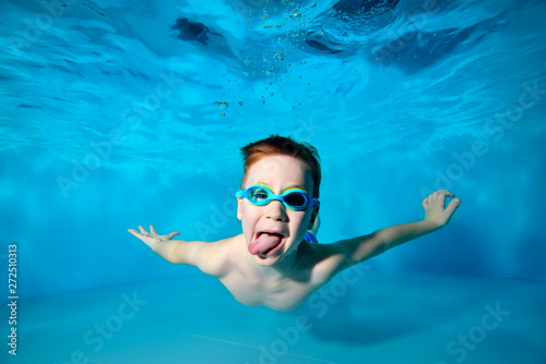 Happy smiling baby, boy, swims underwater in swimming pool in swimming glasses and shows tongue on camera on blue background. Portrait. Underwater photography. Horizontal orientation