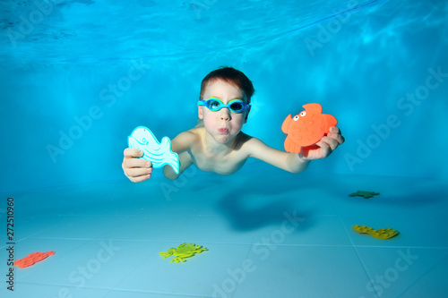 Cute sports baby swims underwater and holds sea toys in his hand. He looks at the camera and smiles. Portrait. Underwater photography. Horizontal orientation