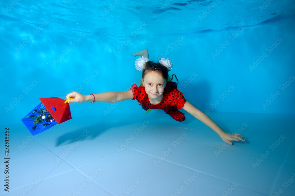 A cute little girl is swimming underwater in the pool with her eyes open at the bottom in a red dress, holding a toy gift in her hand. She smiles and poses for. Portrait. Horizontal view