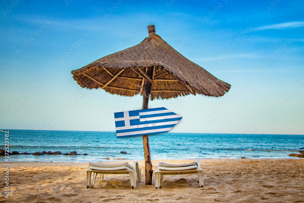 Greek flag on wooden arrow sign. There are two sun loungers and a sun umbrella on the beach. It is a tropical paradise with a clear sea.