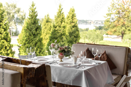 Wedding table setting decorated with fresh flowers in a brass vase. Wedding floristry. Banquet table for guests outdoors with a view of green nature. Bouquet with roses, eustoma and eucalyptus leaves