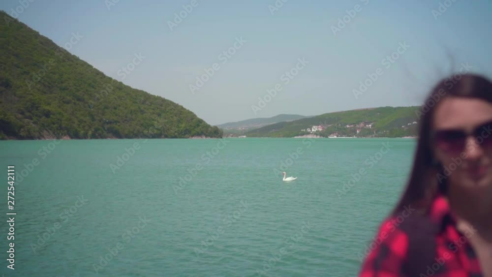 Portrait of a young attractive girl in a red checkered shirt, posing in front of the camera. On a warm day, a girl stands on the shore of a lake, with a white swan in the background. Portrait of a young attractive girl in a red checkered shirt, posing in front of the camera. On a warm day, a girl stands on the shore of a lake, with a white swan in the background.