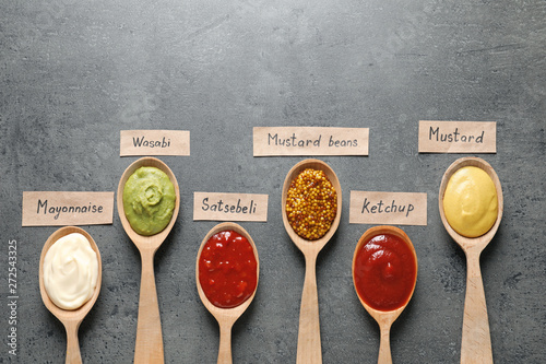 Different sauces in spoons and name tags on gray background, flat lay