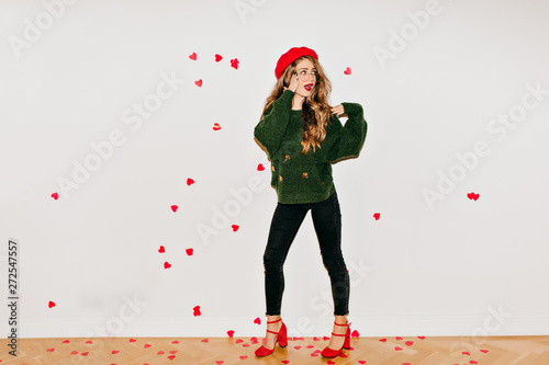 Full-length photo of surprised girl in red sandals and beret standing under heart confetti. Indoor portrait of cheerful brunette lady isolated on white background.